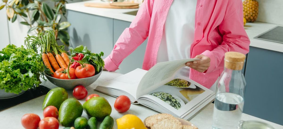 A person browsing a cookbook surrounded by fresh vegetables in a bright kitchen setting.