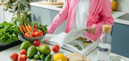 A person browsing a cookbook surrounded by fresh vegetables in a bright kitchen setting.