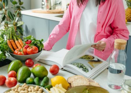 A person browsing a cookbook surrounded by fresh vegetables in a bright kitchen setting.